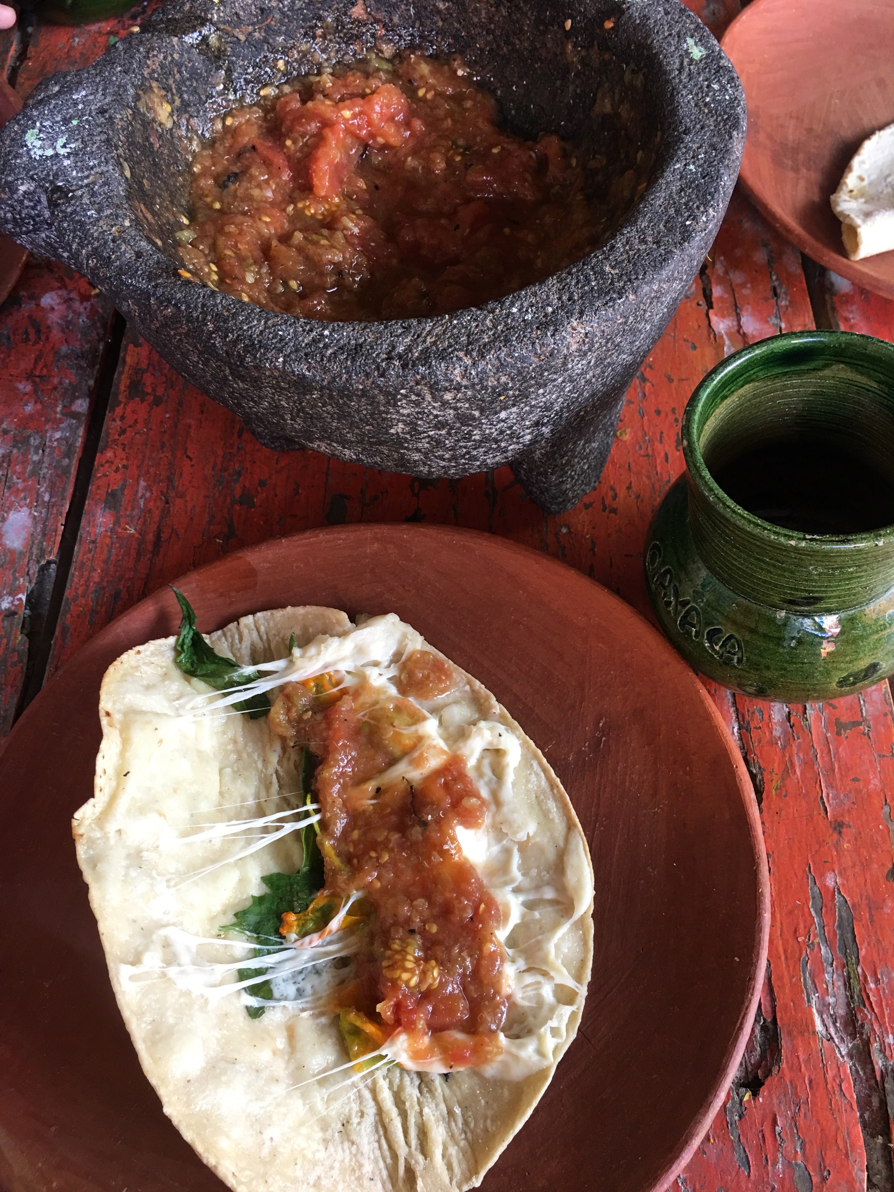 Tortilla with sauce and cheese on a wooden table next to a stone bowl and green cup.