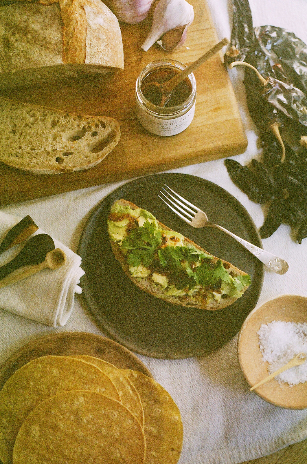 Green plate with a tortilla topped with greens, surrounded by bread, a jar of spread, and salt on a white cloth.